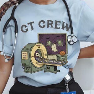 May include: Pale blue t-shirt with the words "CT CREW" in grey. The shirt displays a Halloween-themed illustration of a skeleton in a CT scanner, a ghost, and a pumpkin. A stethoscope is around the neck.