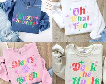 Könnte beinhalten: Vier Sweatshirts mit festlichen Slogans. Ein blaues Sweatshirt hat "Cozy Christmas" in bunten Farben. Ein weißes Sweatshirt sagt "Oh What Fun". Ein rosa Sweatshirt sagt "Merry & Bright". Ein beiges Sweatshirt sagt "Deck the Halls".