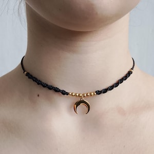 May include: Black macramé choker necklace with gold-coloured beads and a gold crescent moon pendant. The necklace is worn around the neck, a simple, elegant accessory.