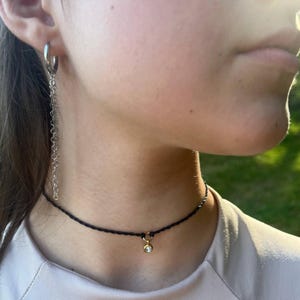 Puede incluir: Un collar gargantilla negro con un pequeño colgante dorado con una piedra preciosa transparente. Un pendiente de cadena plateada cuelga de la oreja. La joya se lleva sobre una camiseta clara.