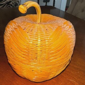May include: A woven basket shaped like an apple, with a curved stem. The basket is made of light orange-yellow wicker, with a lid that fits snugly. The apple basket is a decorative storage item.