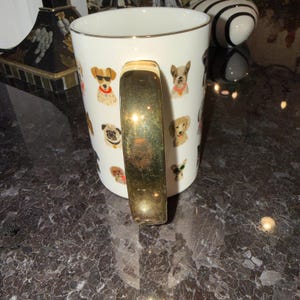Op de afbeelding: Witte keramische mok met een gouden handvat en rand, versierd met kleurrijke illustraties van verschillende hondenrassen. De honden worden afgebeeld met accessoires zoals zonnebrillen en bandana's. Een decoratief item voor hondenliefhebbers.