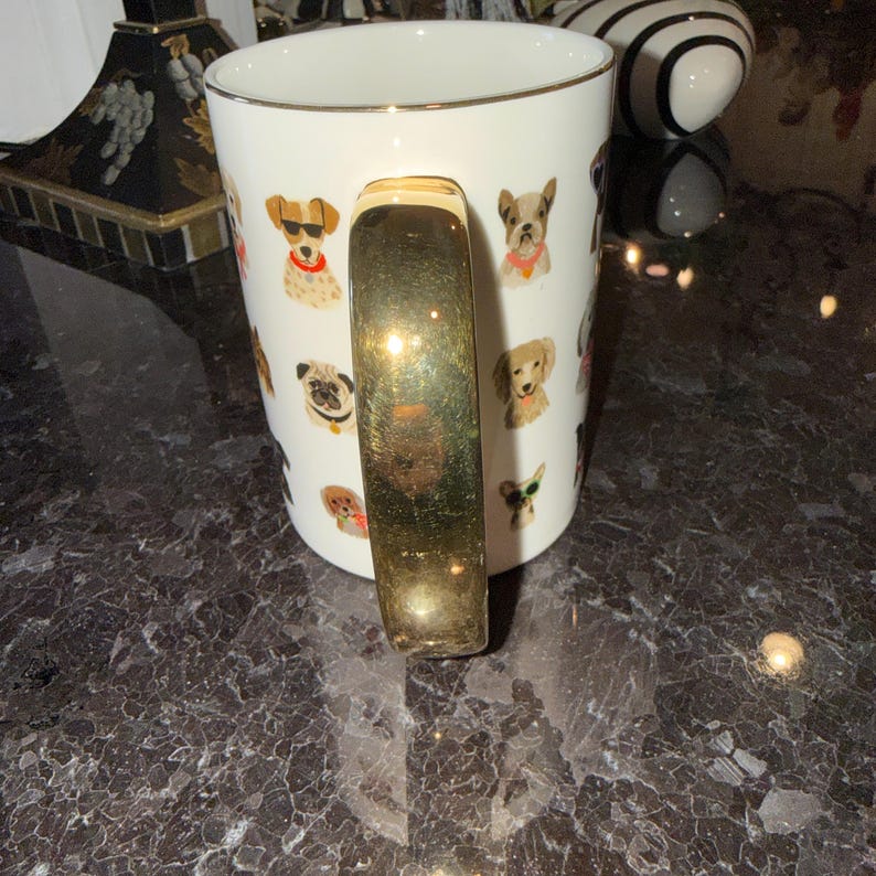 Op de afbeelding: Witte keramische mok met een gouden handvat en rand, versierd met kleurrijke illustraties van verschillende hondenrassen. De honden worden afgebeeld met accessoires zoals zonnebrillen en bandana's. Een decoratief item voor hondenliefhebbers.