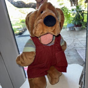 May include: A plush dog toy with a brown head, black nose, and pink tongue. It wears a red corduroy overall with a striped shirt underneath. The toy is soft and cuddly, perfect for children.