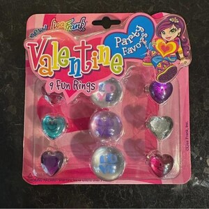 May include: A Lisa Frank Valentine's Day party favour pack with nine fun rings. The rings are heart-shaped and round, with various colours and designs. The packaging is pink with the words "Valentine" and "Party Favours".