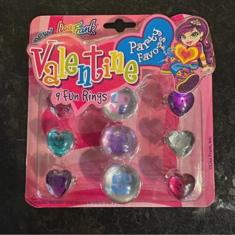 May include: A Lisa Frank Valentine's Day party favour pack with nine fun rings. The rings are heart-shaped and round, with various colours and designs. The packaging is pink with the words "Valentine" and "Party Favours".