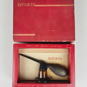 May include: Vintage DeVilbiss nose and throat atomizer in its original red box. The atomizer is black and brown with a glass bottle and a rubber bulb. The box has the brand name and product information printed on it.