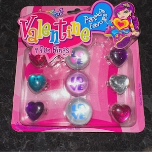 May include: A pack of nine Valentine's Day rings. The rings are heart-shaped and round, some with the word "LOVE". The pink packaging features the word "Valentine" in colourful letters and a cartoon girl.