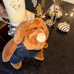 May include: A plush brown dog toy wearing blue denim shorts. The dog has a white nose, red eyes, and large floppy ears. The toy is positioned on a dark surface, with other decorative items in the background.