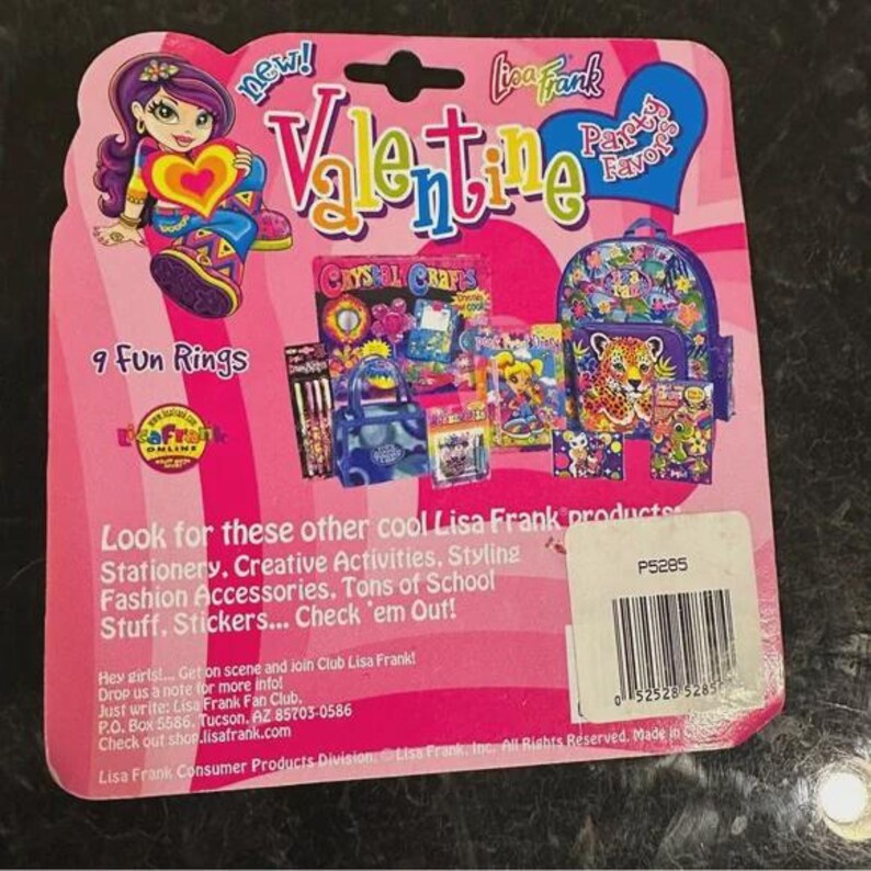 May include: A pink Lisa Frank Valentine's Day party favours package. The package features a cartoon girl holding a heart, the words "Valentine" and "Party Favours", and images of school supplies and a backpack. The package includes 9 fun rings.
