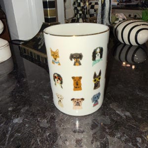 Op de afbeelding: Witte keramische mok met een gouden rand, versierd met kleurrijke illustraties van verschillende hondenrassen. De honden zijn afgebeeld met accessoires zoals zonnebrillen en bandana's. De mok is cilindrisch en staat op een donker, getextureerd oppervlak.