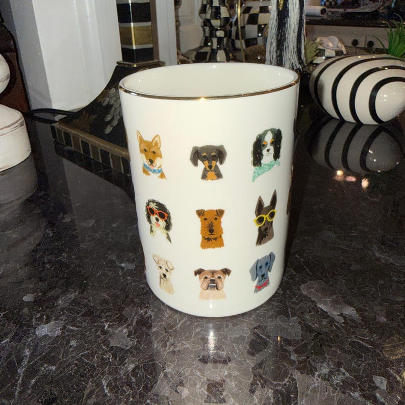 Op de afbeelding: Witte keramische mok met een gouden rand, versierd met kleurrijke illustraties van verschillende hondenrassen. De honden zijn afgebeeld met accessoires zoals zonnebrillen en bandana's. De mok is cilindrisch en staat op een donker, getextureerd oppervlak.