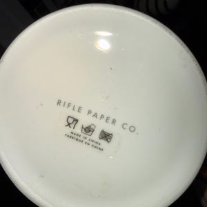 Op de afbeelding: Een close-up van de bodem van een witte keramische mok. De tekst "RIFLE PAPER CO." is in grijs gedrukt, samen met verzorgingssymbolen en de tekst "MADE IN CHINA FABRIQU&Eacute; EN CHINE".