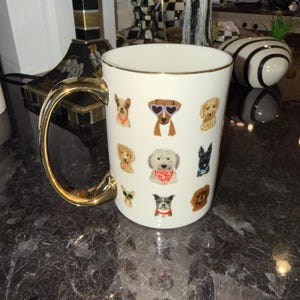 Op de afbeelding: Witte keramische mok met een gouden handvat en rand. De mok is versierd met kleurrijke illustraties van verschillende hondenrassen, elk met accessoires zoals zonnebrillen of bandana's. Een decoratief item voor hondenliefhebbers.