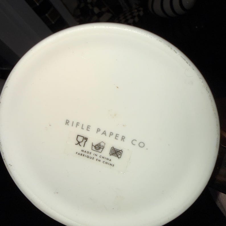 Op de afbeelding: Een wit keramisch object met de tekst "RIFLE PAPER CO." in grijs gedrukt. Daaronder een klein label met symbolen en de tekst "MADE IN CHINA FABRIQUE EN CHINE."