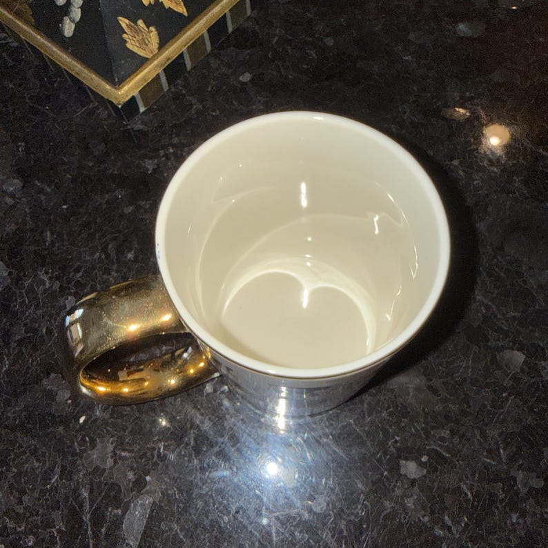 Op de afbeelding: Een witte keramische mok met een gouden handvat en rand. De mok is leeg en staat op een donker, gespikkeld oppervlak. Een decoratieve doos met gouden accenten is gedeeltelijk zichtbaar.