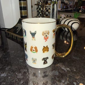 Op de afbeelding: Witte keramische mok met een gouden handvat en rand, versierd met kleurrijke illustraties van verschillende hondenrassen. De mok heeft een standaardformaat voor koffie of thee.