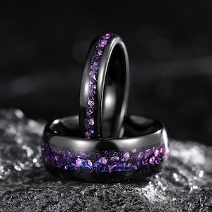 May include: Two black rings with a band of purple and blue crushed stone. One ring is upright, the other is lying down. The rings are set against a dark, textured background, creating a striking contrast.