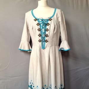 May include: A white linen dress with a blue and silver trim. The dress has a lace-up bodice with silver buttons and a floral embroidered hem.