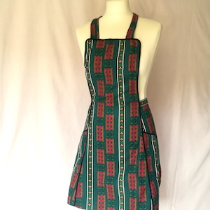 May include: A green apron with a floral pattern in red and white, displayed on a mannequin. The apron has black trim, a bib, and two pockets. The straps are also green with black trim.