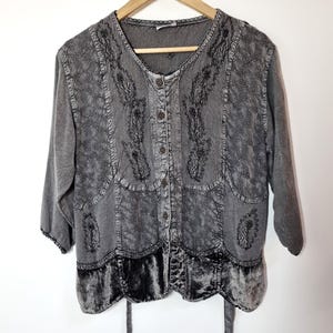 Könnte beinhalten: Graues Hemd zum Knöpfen mit Dreiviertelärmeln, hängt an einem Holzbügel. Das Hemd hat aufwendige schwarze Stickereien und einen Saum aus Samt. Der Stoff hat ein strukturiertes, ausgewaschenes Aussehen.