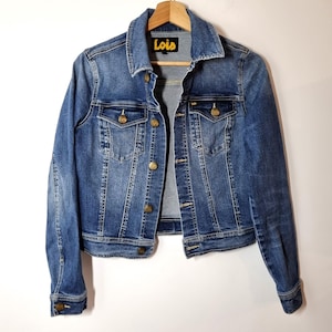 Könnte beinhalten: Blaue Jeansjacke mit klassischem Design, mit Kragen, Knopfleiste und zwei Brusttaschen. Die Jacke hat goldfarbene Knöpfe und der Markenname "Lois" ist auf dem Etikett sichtbar. Die Jacke hängt an einem Holzkleiderbügel.