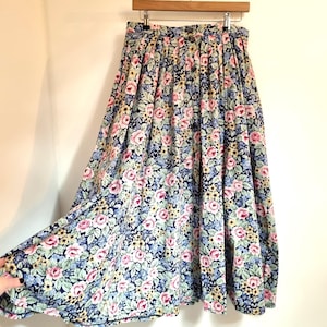 Puede incluir: Falda larga con estampado floral, con fondo azul marino, adornada con rosas rosas, detalles amarillos y hojas azul claro. La falda está colgada de una percha de madera. El diseño es plisado.