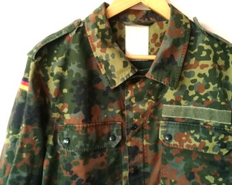 Puede incluir: Una camisa de camuflaje verde, marrón y negro con cierre de botones y un parche en la manga. La camisa tiene una bandera alemana en la manga.