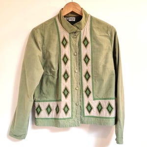 May include: A pale green jacket with a stand-up collar and long sleeves. The jacket has a cream-coloured panel down the front, decorated with green diamond-shaped embroidery. It fastens with buttons and is on a wooden hanger.