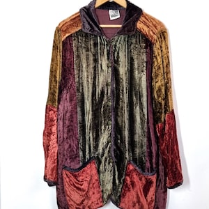 Op de afbeelding: Een veelkleurige fluwelen jas met capuchon en twee voorzakken. De jas heeft panelen van bruin, bordeauxrood, olijfgroen en roestkleurig fluweel. Het hangt aan een houten kleerhanger tegen een witte achtergrond.