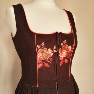 K&ouml;nnte beinhalten: Ein braunes, &auml;rmelloses Kleid mit rotem Besatz und gestickten Blumenmotiven. Das Kleid hat einen Rei&szlig;verschluss vorne.