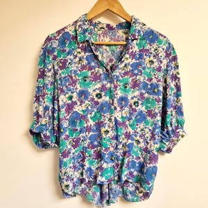 May include: A white button-up shirt with a floral print in shades of blue, purple, and green. The shirt has a collar and short sleeves.