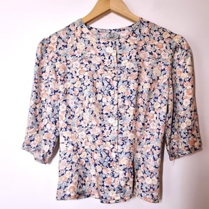 Op de afbeelding: Een vintage bloemenblouse met knopen. De blouse heeft een marineblauwe achtergrond met een patroon van kleine bloemen in tinten perzik, crème en lichtblauw. De mouwen zijn driekwart lang.