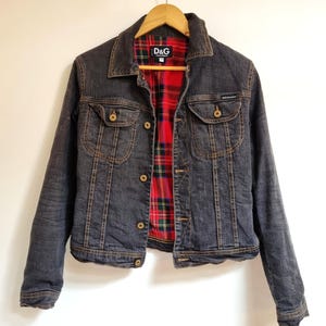 May include: A dark gray denim jacket with a red and black plaid lining. The jacket has two chest pockets, button closures, and the D&G logo on the inner label. The jacket is hanging on a wooden hanger.