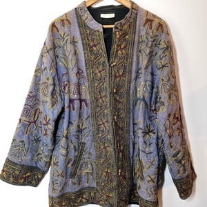 Può includere: Giacca azzurra con ricami intricati e colletto alla coreana. La giacca presenta un motivo dettagliato di disegni floreali e geometrici in oro, marrone e viola. Ha una fodera nera e chiusure a toggle in legno. La giacca è appesa a una gruccia di legno.