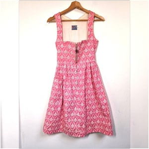 May include: A pink dress with a white floral pattern, square neckline, and a zipper down the front. The dress has wide straps and a fitted bodice that flares into a pleated skirt. It hangs on a wooden hanger.