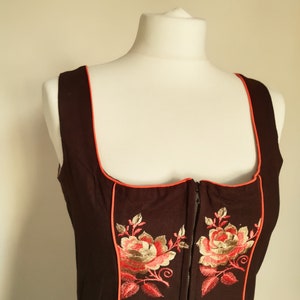 K&ouml;nnte beinhalten: Ein schwarzes Dirndl-Mieder mit rotem Besatz und gestickten Blumenmotiven. Das Mieder hat einen Rei&szlig;verschluss vorne.
