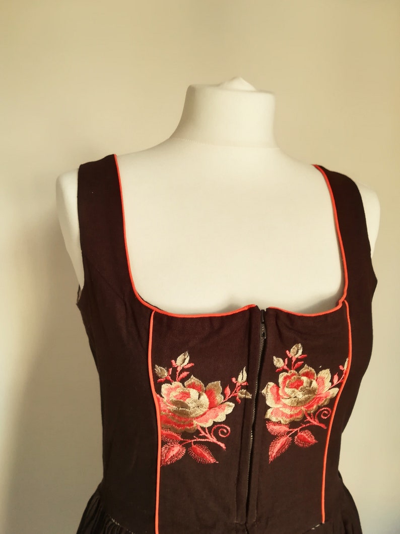 K&ouml;nnte beinhalten: Ein schwarzes Dirndl-Mieder mit rotem Besatz und gestickten Blumenmotiven. Das Mieder hat einen Rei&szlig;verschluss vorne.