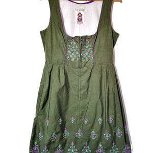 May include: An olive green sleeveless dress featuring a square neckline, a front zipper closure, and purple and light green floral embroidery along the bodice and hem. The interior lining is white with a small logo tag at the center neckline.