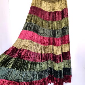 May include: A floor-length skirt featuring a crushed velvet texture with horizontal stripes in alternating shades of olive green, deep burgundy, and dark forest green. The garment has an elasticated waistband and hangs from a wooden hanger.