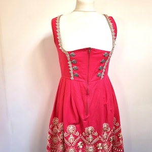 May include: A bright pink dress with a fitted bodice and a flared skirt. The dress features decorative silver trim along the straps and bodice, with ornate silver buttons. The skirt has an embroidered floral pattern in gold and white.