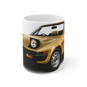 May include: A white ceramic mug featuring a detailed graphic of a gold-coloured sports car with pop-up headlights, a black front bumper, and silver wheels. The car is depicted from a front-quarter angle, showcasing its sleek design and windscreen.