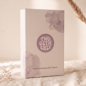 May include: A rectangular white box featuring a central purple circle with the text TWOGETHERLY printed in white. The box is decorated with soft, light purple watercolor-style floral shapes and includes the text Das Kartenset für Paare.