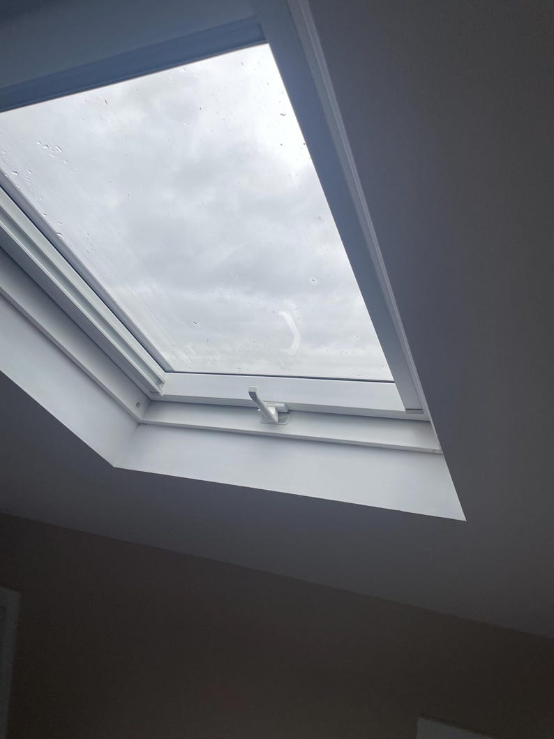 Puede incluir: Un tragaluz rectangular blanco instalado en un techo, inclinado hacia el espectador. El cristal revela un cielo nublado y cubierto. Se ve una manija plateada en el marco. Las paredes circundantes est&aacute;n pintadas de un gris neutro.