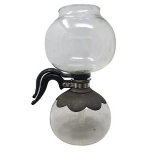 May include: A clear glass siphon coffee maker with a round top and bottom chamber. The bottom chamber has a decorative metal base. It features black handles and a glass tube connecting the chambers. A vintage coffee brewing device.