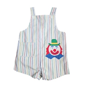 May include: A white romper featuring vertical stripes in red, yellow, green, and blue. The garment has two shoulder straps with white buttons, a side pocket, and a large embroidered clown face appliqué with a green hat and blue bow tie.