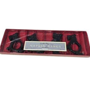 May include: Set of four black Scottie dog napkin rings in a red box. Each ring is shaped like a Scottie dog with a circular opening for a napkin. The box has a label that reads "Scottie Dog Napkin Rings."