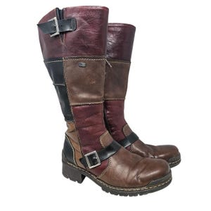 May include: A pair of knee-high leather boots in a patchwork design. The boots feature a mix of brown, burgundy, and black panels, with buckle and zipper details. The soles are lugged, and the boots have a stacked heel.
