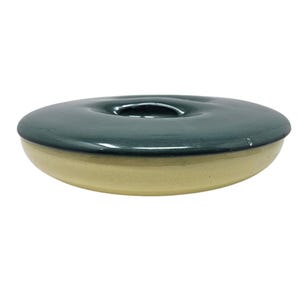 May include: A ceramic, two-toned serving dish with a dark teal lid and a light tan base. The lid has a circular indentation in the center. The dish is round and appears to be handmade.