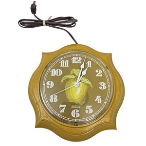 May include: A vintage Sunbeam wall clock with a gold-colored, scalloped frame. The clock face is olive green with white numerals and hands. A yellow apple graphic is in the center. The clock has a brown power cord.